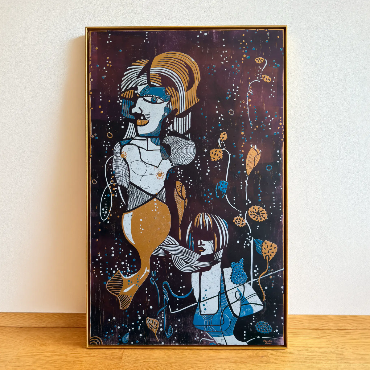 The linocut Underwater Love, framed in a gold shadowbox frame, stands on a wooden floor and is leaning against the beige wall. It depicts two stylized figures among aquatic plants and bubbles.