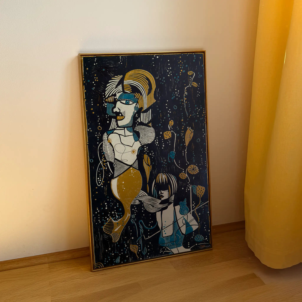 The linocut Underwater Love, framed in a gold shadow-joint frame, stands on a wooden floor and is leaning against the beige wall. It depicts two stylized figures among aquatic plants and bubbles.