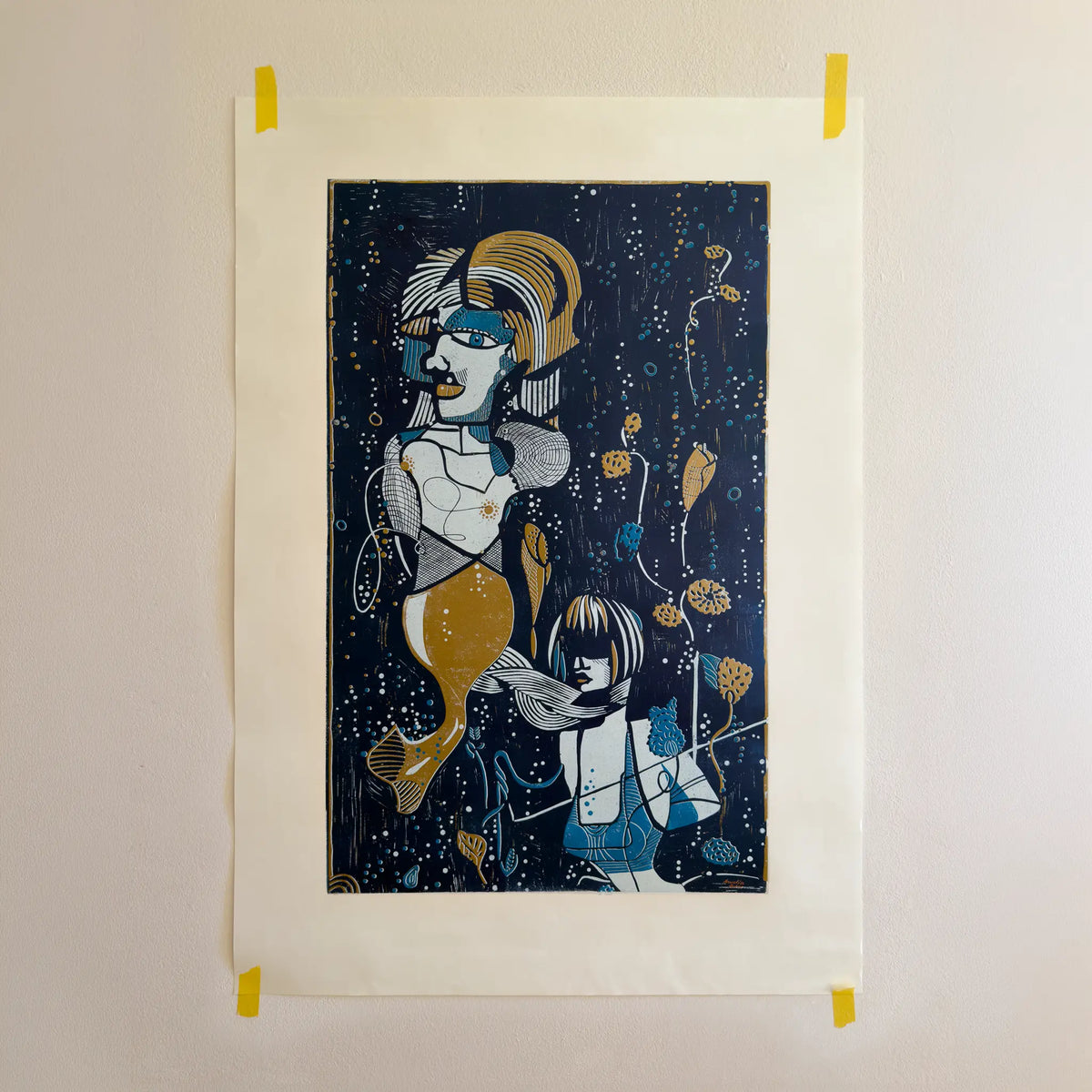 The linocut “Underwater Love” in gold and blue, attached to a beige wall with yellow tape.