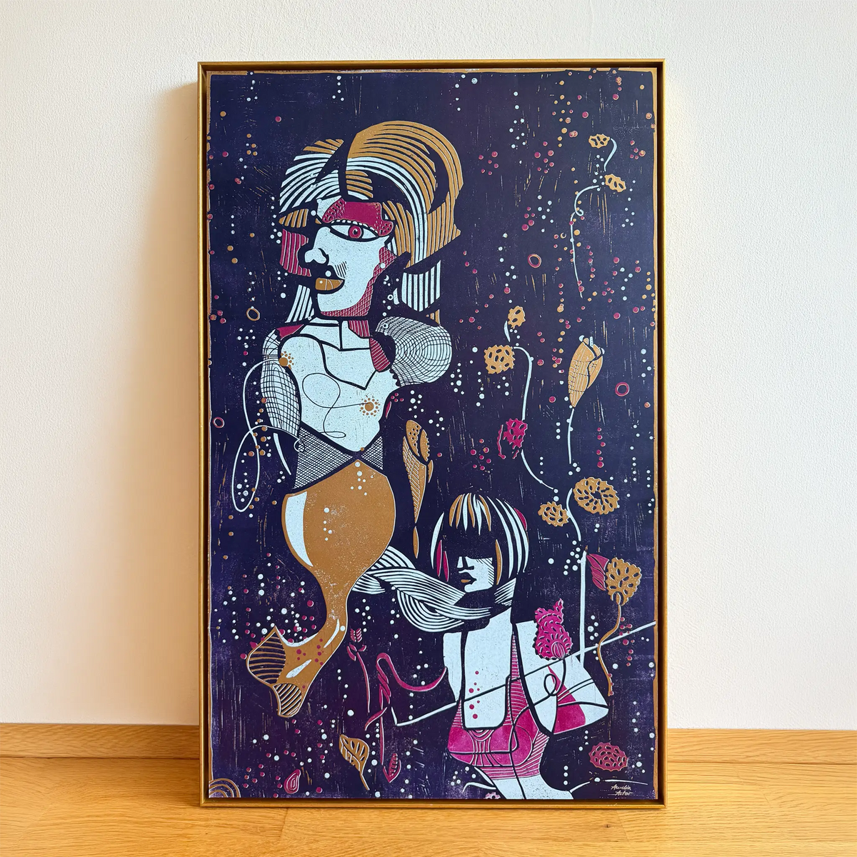 The linocut Underwater Love, framed in a gold shadow-joint frame, stands on a wooden floor and is leaning against the beige wall. It depicts two stylized figures among aquatic plants and bubbles.