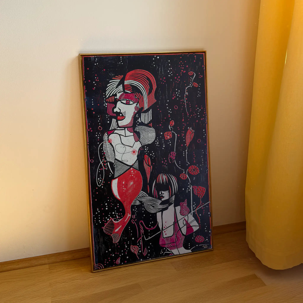 The linocut Underwater Love, framed in a gold shadow-joint frame, stands on a wooden floor and is leaning against the beige wall. It depicts two stylized figures among aquatic plants and bubbles.