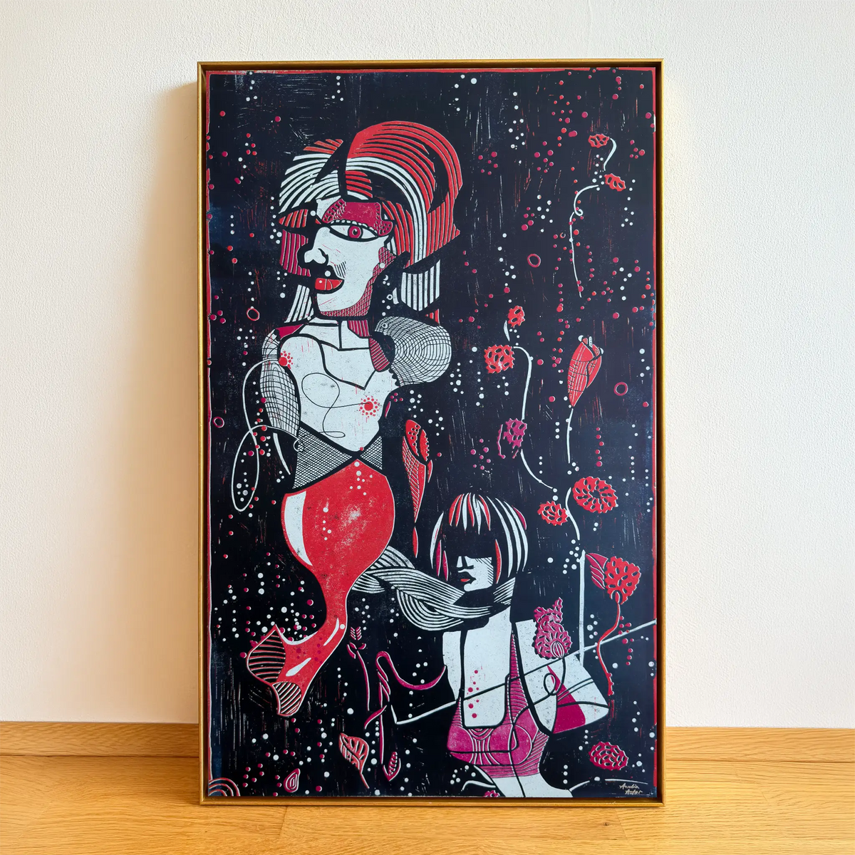 The linocut Underwater Love, framed in a gold shadow-joint frame, stands on a wooden floor and is leaning against the beige wall. It depicts two stylized figures among aquatic plants and bubbles.