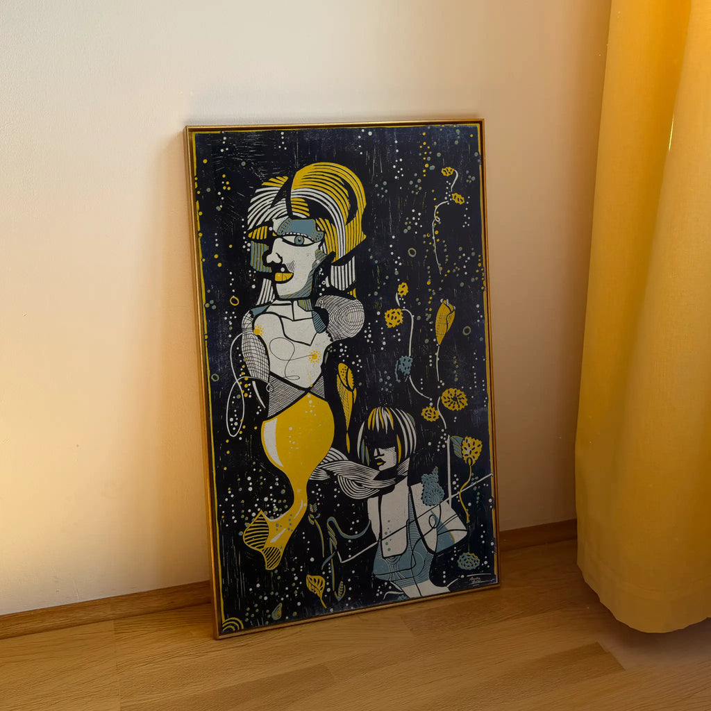 The linocut Underwater Love, framed in a gold shadow-joint frame, stands on a wooden floor and is leaning against the beige wall. It depicts two stylized figures among aquatic plants and bubbles.