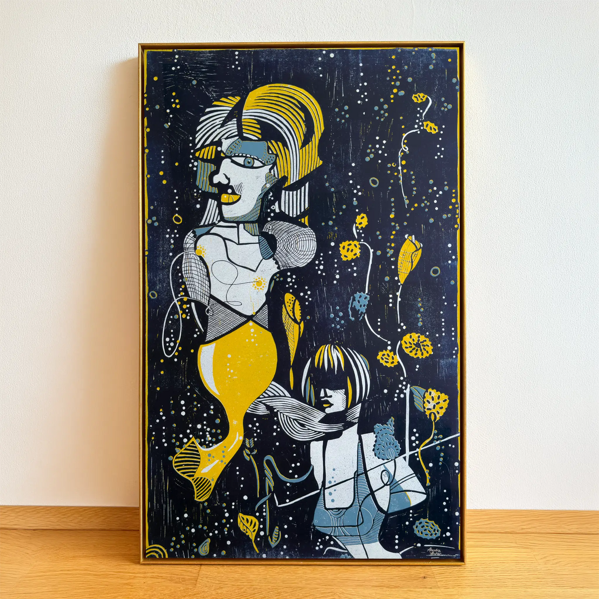 The linocut Underwater Love, framed in a gold shadow-joint frame, stands on a wooden floor and is leaning against the beige wall. It depicts two stylized figures among aquatic plants and bubbles.
